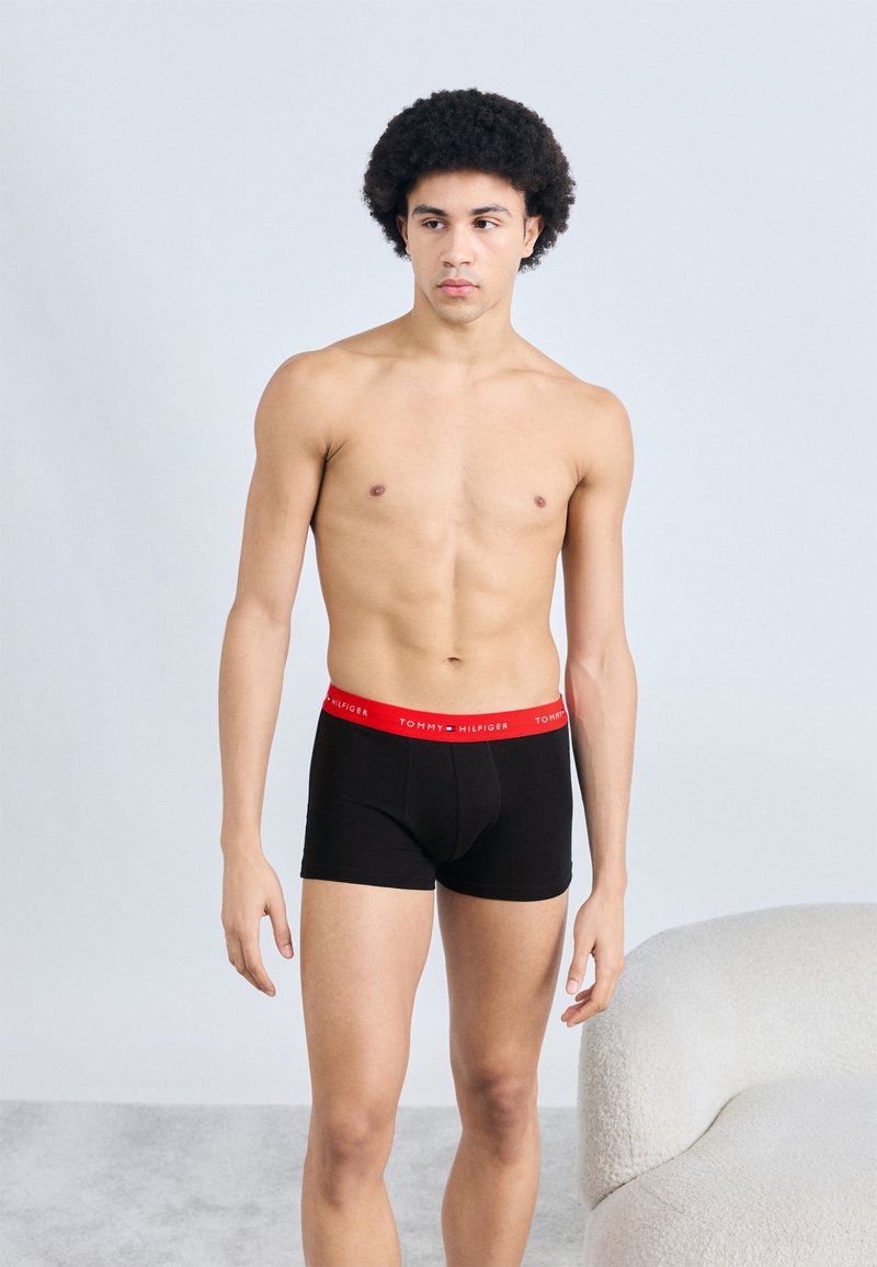 Μαύρα μποξεράκια με κόκκινη μέση και το λογότυπο "TOMMY HILFIGER". Το μοντέλο στέκεται, παρουσιάζοντας τη φόρμα και το κόψιμο. Ουδέτερο φόντο.