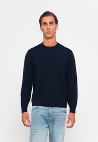 Pulover bleumarin cu guler rotund dintr-un material moale, cu mâneci lungi și guler reiat. Asortat cu blugi deschiși la culoare, deteriorați.