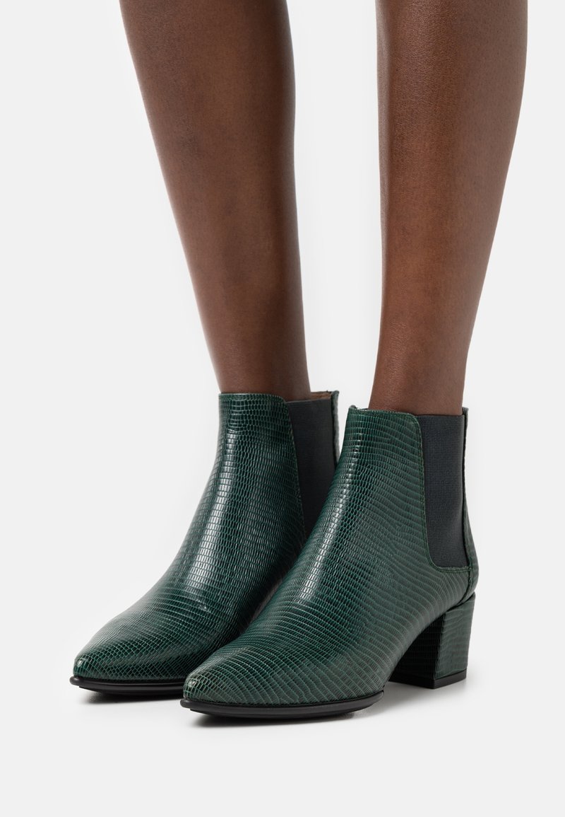 Bottes de cheville texturées vert foncé avec talons carrés portées sur des jambes de peau foncée, présentées sur un fond blanc uni.