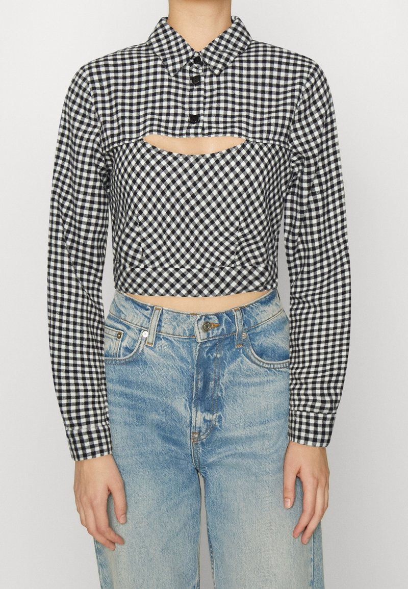 Chemise à manches longues écourtée à carreaux noirs et blancs avec une découpe au-dessus d'un jean taille haute bleu clair portée par une personne debout.