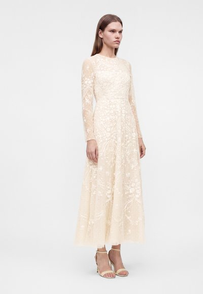 Jeune femme debout portant une robe en dentelle fleurie crème à manches longues avec des manches transparentes et des sandales à talons beiges sur un fond uni.