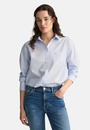 Chemise button-up bleu clair en coton avec une poche, associée à un jean bleu. La chemise présente des manches longues et un col classique.