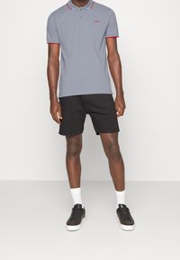 Polo gris avec des accents rouges, patte de boutonnage à trois boutons, logo sur la poitrine, associé à un short noir et des baskets noires. Chaussettes blanches visibles.