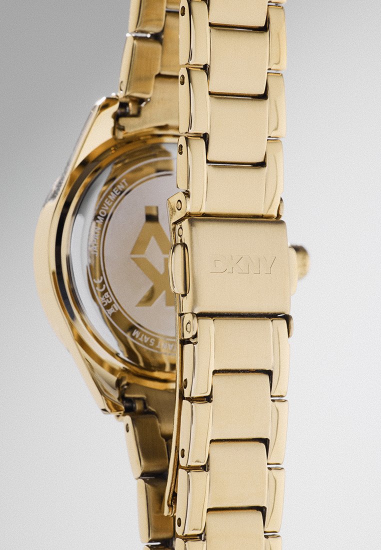 Gouden metalen polshorloge met een schakelarmband, ronde wijzerplaat en gegraveerd logo op de sluiting. Door de transparante achterkant zijn de details van het uurwerk zichtbaar.