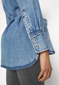 Denim srajca v svetlo modri barvi, klasičnim ovratnikom, z gumbnimi manšeti in ukrivljenim robom. Tkanina prikazuje subtilno teksturo in bledenje.