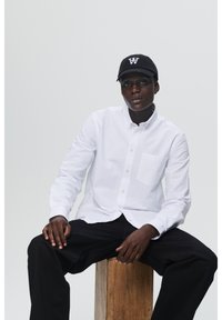 Camisa blanca con botones y un bolsillo en el pecho, combinada con pantalones negros y una gorra negra con un logo blanco, sentado sobre una superficie de madera.