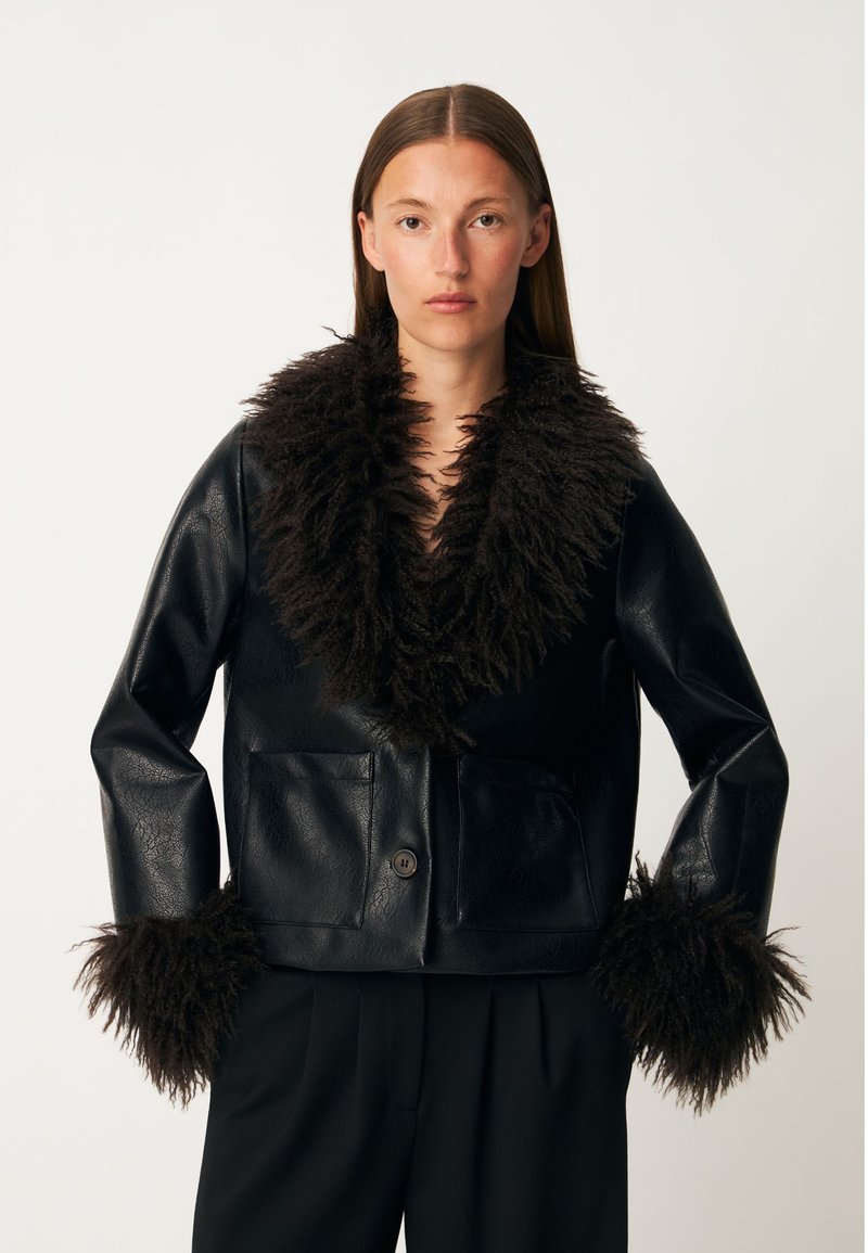 Femme portant une veste en cuir noire avec un grand col et poignets en fourrure duveteuse, debout les mains sur les hanches devant un fond uni.