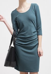 Robe bleu sarcelle longueur genou avec manches trois-quarts ; présente un col rond échancré et des détails froncés à la taille. Associée à un sac à main noir.