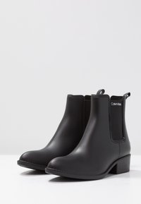 Pair of black matte ankle boots with low heels and elastic side panels, featuring a pull tab and Calvin Klein logo on the side.