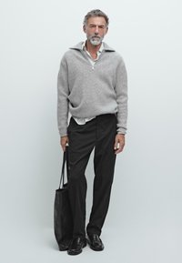 Volwassen man in grijze zip-neksweater, witte shirt, zwarte broek, zwarte instappers, met een zwarte leren tas, staand tegen een effen witte achtergrond.