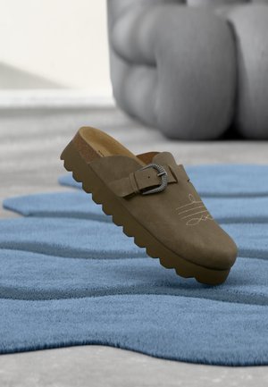 Zueco marrón de ante sin cordones con hebilla de metal y diseño ondulado cosido, reposando sobre una alfombra azul texturizada en el interior.