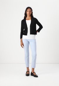 Zwarte cropped cardigan met driekwartmouwen, gedragen over een witte top, gecombineerd met lichtblauwe slim-fit broek en zwarte schoenen.