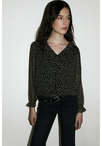 Zwarte blouse met klein wit bloemmotief, V-hals, knoopsluiting aan de voorkant en doorzichtige lange mouwen. Gecombineerd met een donkere spijkerbroek en een zwarte riem.