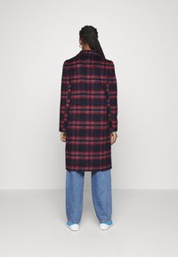 Nainen, jolla on letitetyt hiukset, pukeutuneena polvimittaiseen tummansiniseen takkiin, jossa on punaisia ruutuja, vaaleansinisiin farkkuihin ja valkoisiin lenkkikenkihin, joissa on sinisiä yksityiskohtia, kääntymässä pois.