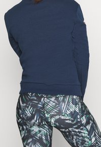 Persoon die een marineblauwe sweatshirt en zwarte leggings draagt met groene en witte abstracte bladerenpatronen, gezien van achteren.