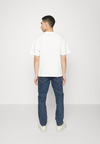 Camiseta de algodón blanca con mangas cortas, de corte relajado, vista desde la parte trasera, combinada con pantalones vaqueros azules y zapatillas blancas.