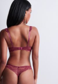 Femme avec de longs cheveux noirs portant de la lingerie en dentelle bourgogne, vue de dos sur un fond clair et uni.