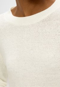 Maglione bianco lavorato a maglia con scollatura a coste, che mette in evidenza un tessuto testurizzato e sottili variazioni nel colore del filo. Vista ravvicinata.