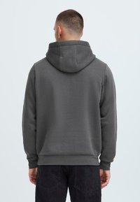 Felpa grigia con cappuccio, dal texture morbido, con cappuccio a doppio strato e polsini a costine. Indossata con pantaloni scuri, mostrando una vestibilità comoda.