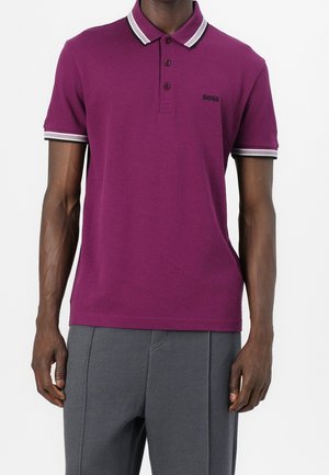 Homme portant un polo à manches courtes violet avec un col et des bordures de manches rayés noirs et blancs, associé à un pantalon gris texturé.