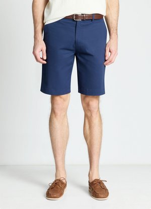 Uomo con pantaloncini blu fino al ginocchio, cintura in pelle marrone e mocassini marroni con lacci, in piedi su uno sfondo bianco semplice.