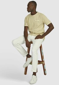 Beige katoenen t-shirt met een borstzak, gecombineerd met witte denim broeken en witte sneakers. Model zittend op een houten kruk.