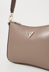 Beige syntetisk handväska med böjd form, guldfärgad hårdvara och en framträdande triangel-logotyp. Har ett blixtlås och ett kort handtag.