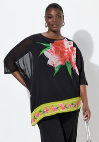 Blusa negra transparente con un gran estampado floral en rosa y blanco, que presenta un acento en el dobladillo verde y rosa. Diseño suelto y fluido.