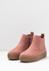 Par de botas de ante rosa empolvado hasta el tobillo con cremallera lateral, panel elástico, tira de agarre y suelas de goma marrón gruesas sobre una superficie blanca.