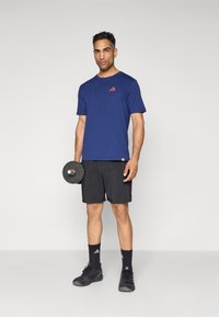Man in een blauw T-shirt, zwarte shorts en zwarte sneakers staat met een enkele dumbbell naast zich tegen een effen achtergrond.