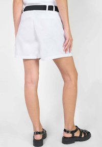 Pantalones cortos blancos con un cinturón negro, bolsillos frontales y un corte clásico. El material parece ligero y tiene una textura suave. Las sandalias negras complementan el conjunto.