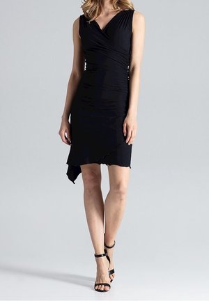 Femme portant une robe portefeuille asymétrique noire sans manches et des sandales à talons hauts noires à bride à la cheville, debout devant un fond gris.