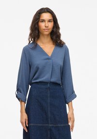 Blaue, gemusterte Bluse mit einem Kreuz-Frontdesign und dreiviertellangen Ärmeln, kombiniert mit einem hoch taillierten Jeansrock mit sichtbaren Nähten.