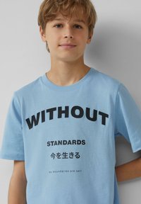 Hellblaue Baumwoll-T-Shirt mit kurzen Ärmeln. Verfügt über fettgedruckten schwarzen Text: "OHNE STANDARDS" und japanische Schriftzeichen, mit einem kleinen Slogan darunter.