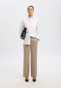 Camicia bianca con bottoni, pantaloni beige su misura, tacchi a punta neri, in mano una borsa di pelle nera con dettaglio a fiocco. Tessuti lisci.