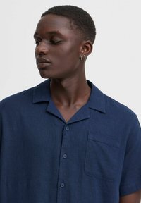Camisa de manga corta azul marino hecha de tejido texturizado, con cuello tipo punta, cierre de botones en el frente y un bolsillo en el pecho.