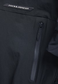 Veste noire avec une poche zippée dotée d'une tirette texturée et d'un logo discret sur la poitrine. Tissu lisse et léger.