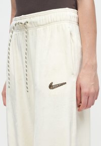 Személy fehér bolyhos melegítőnadrágot visel, fekete-fehér zsinórral és leopárdmintás Nike logóval a combján, keze oldalt lazán lóg.