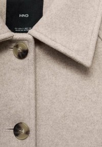 Cappotto in lana beige con un colletto deciso, due grandi chiusure a bottoni e una consistenza morbida. Etichetta visibile all'interno con dettagli del marchio.