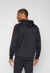 Schwarzer Hoodie mit taillierter Passform, strukturiertem Stoff und Kapuze. Mit roten Streifen entlang der Schultern. Modell von hinten gezeigt.