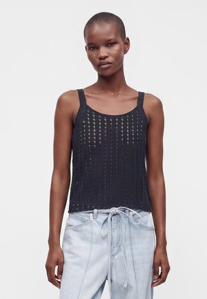 Kvinde med kort hår iført en ærmeløs sort mesh-top og lyseblå jeans med bælte, der bindes foran, står mod en ensfarvet baggrund.