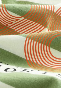 Grüner und weißer Stoff mit gebogenen orangefarbenen Linien, die geometrische Muster bilden, und einem Teil schwarzen Textes auf einer weichen, gefalteten Oberfläche.