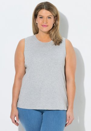 Frau mit hellbraunen Haaren, die silberne Creolen, ein ärmelloses graues Oberteil und blaue Jeans trägt, steht vor einem hellen Hintergrund.