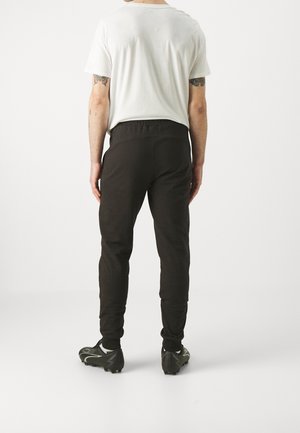 Mężczyzna stojący tyłem, ubrany w białą koszulkę, czarne spodnie dresowe i czarne sportowe korki na jednolitym tle.