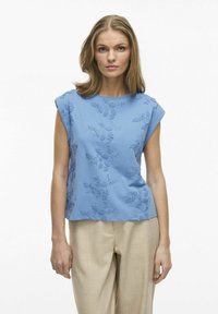 Jasnoniebieska bluzka z krótkim rękawem z delikatnym haftem w kwiaty. Luźny krój i okrągły dekolt, w połączeniu z beżowymi spodniami. Gładka faktura.