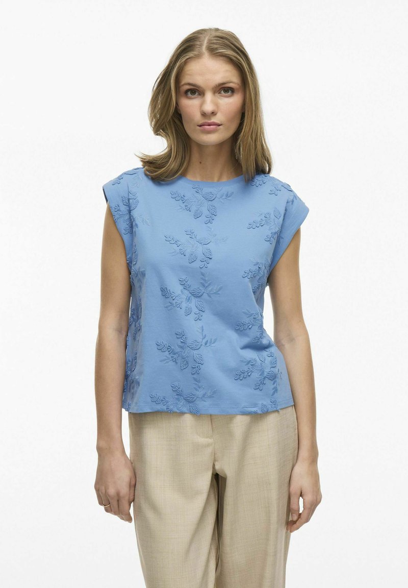 Jasnoniebieska bluzka z krótkim rękawem z delikatnym haftem w kwiaty. Luźny krój i okrągły dekolt, w połączeniu z beżowymi spodniami. Gładka faktura.
