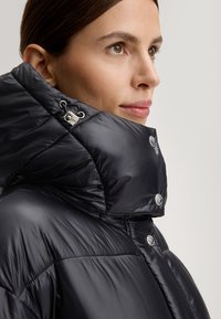 Kobieta w czarnej błyszczącej puchowej kurtce z wysokim kołnierzem i srebrnymi zatrzaskami, patrząca w prawo na jednolitym tle.