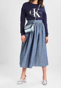 Marinblå sweatshirt med vit Calvin Klein-logotyp, blå kjol i chambray med hög midja, silverfärgade blockklackade skor och liten ljusblå handväska.