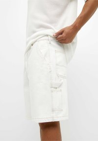Shorts de mezclilla blanca con un corte relajado, bolsillos laterales y costuras en beige contrastantes. La textura es suave y ligeramente rígida.