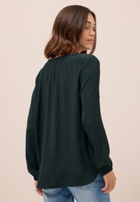 Blusa lungo verde scuro a maniche lunghe realizzata in tessuto leggero. Presenta un colletto arricciato e polsini elastici. Design fluente con orlo arrotondato.